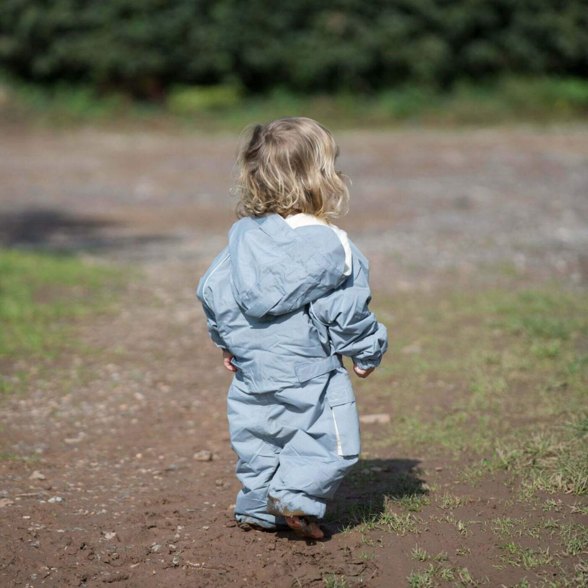 Back view of toddler walking along a muddy path wearing a grey fleece lined waterproof suit with hood up in natural light.