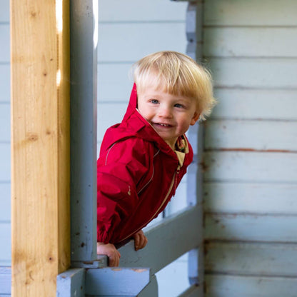 Blonde toddler smiling and leaning on a wooden railing, wearing a bright red waterproof fleece-lined suit, with a light blue background.