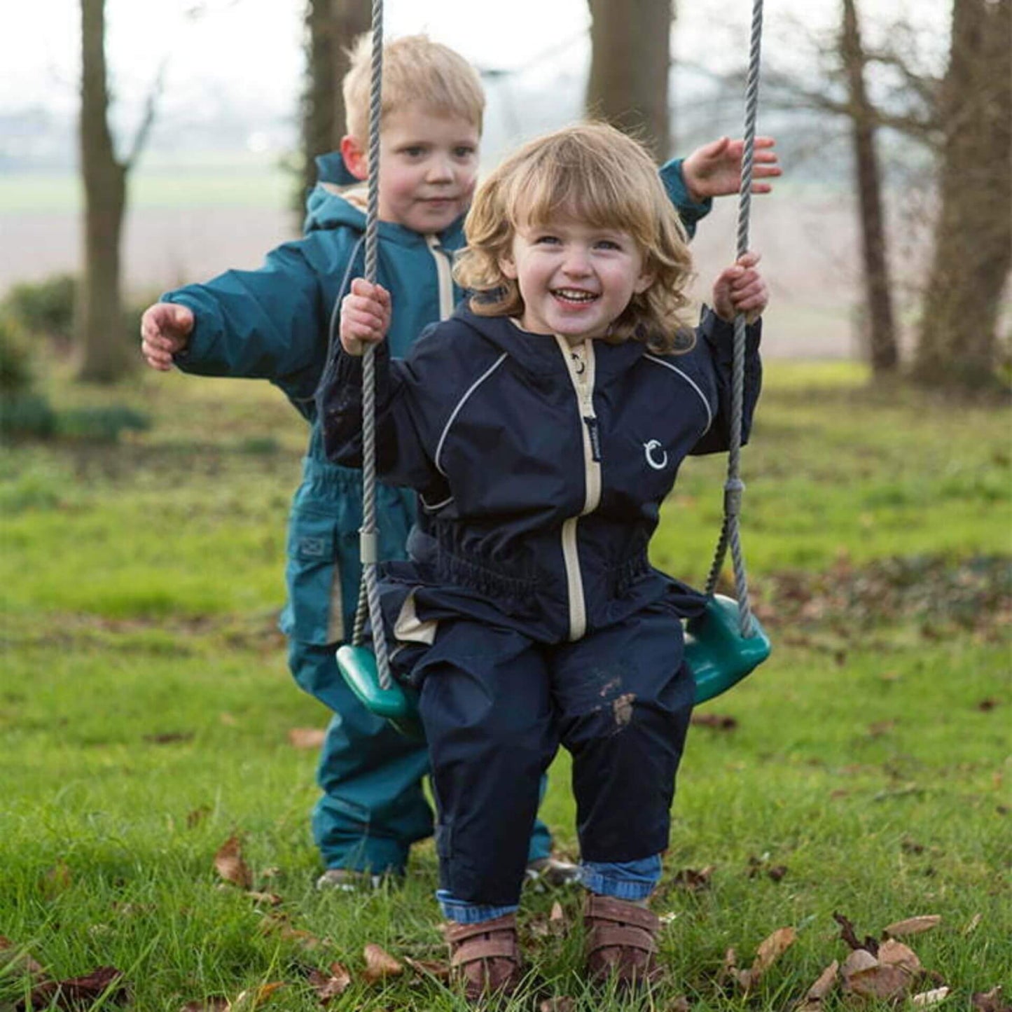 Child sitting on a swing wearing a navy waterproof fleece lined suit with beige zip, smiling while another child stands behind.