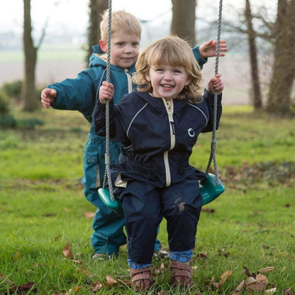 Child sitting on a swing wearing a navy waterproof fleece lined suit with beige zip, smiling while another child stands behind.