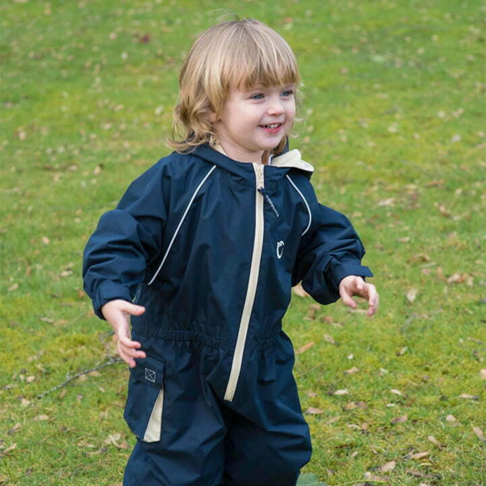 Smiling toddler playing outdoors on grass wearing a navy waterproof fleece lined suit with beige zip and reflective piping.