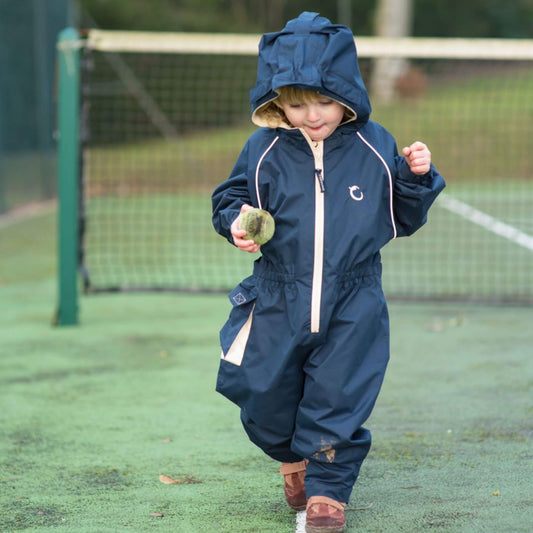 Toddler walking on a tennis court wearing a navy waterproof fleece lined suit with beige zip and hood, holding a tennis ball.