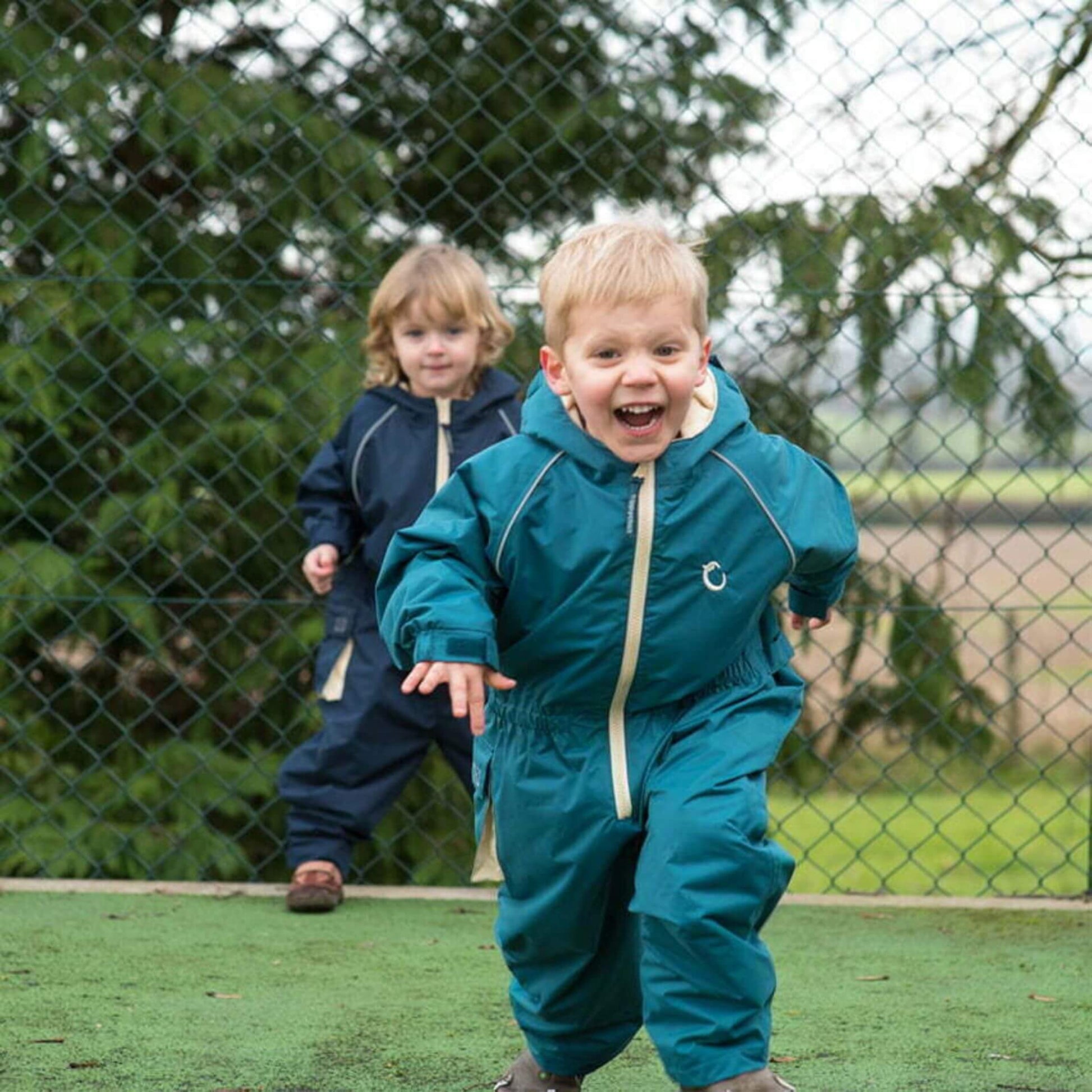 Two children playing and running outdoors, with the child in the foreground wearing a peacock green waterproof fleece-lined suit, and the child in the background wearing a midnight blue suit.