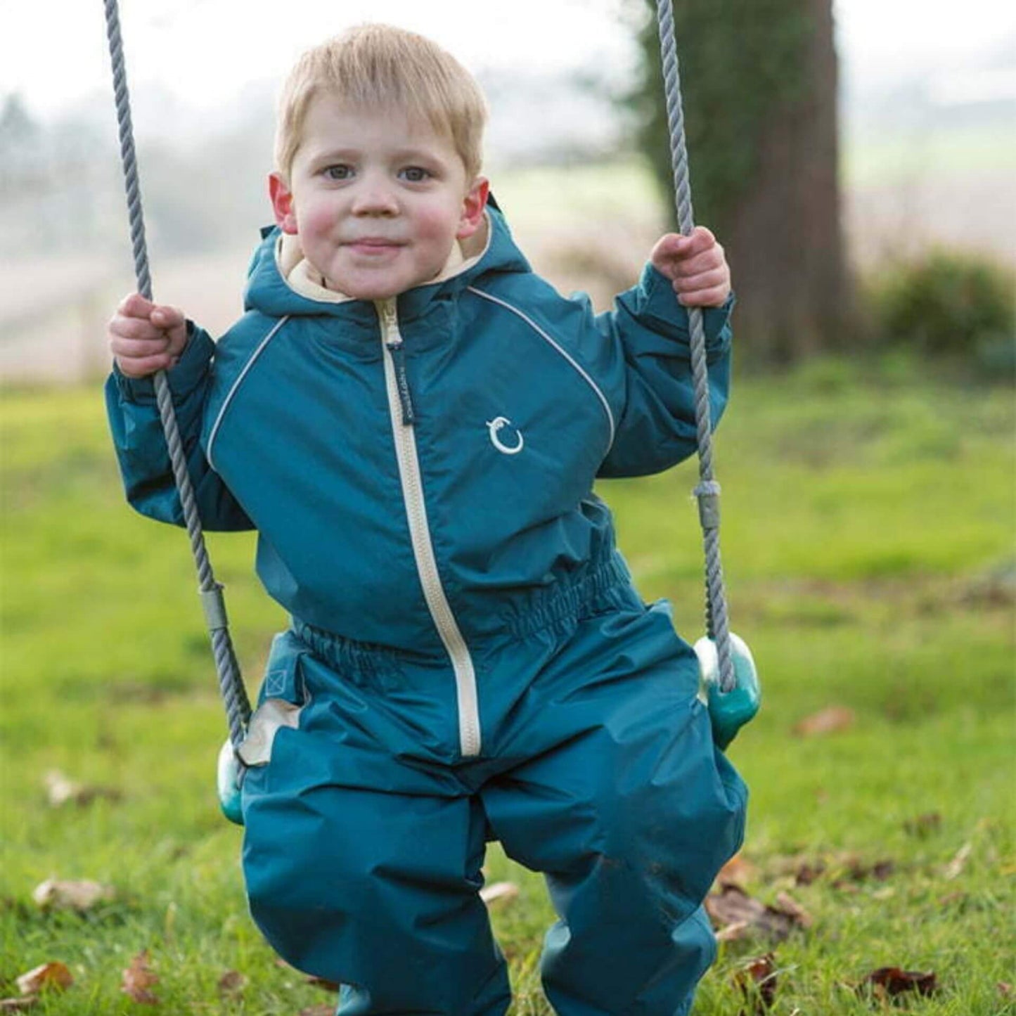 Child wearing a green waterproof fleece-lined suit, sitting on a swing in an outdoor setting with a grassy background.