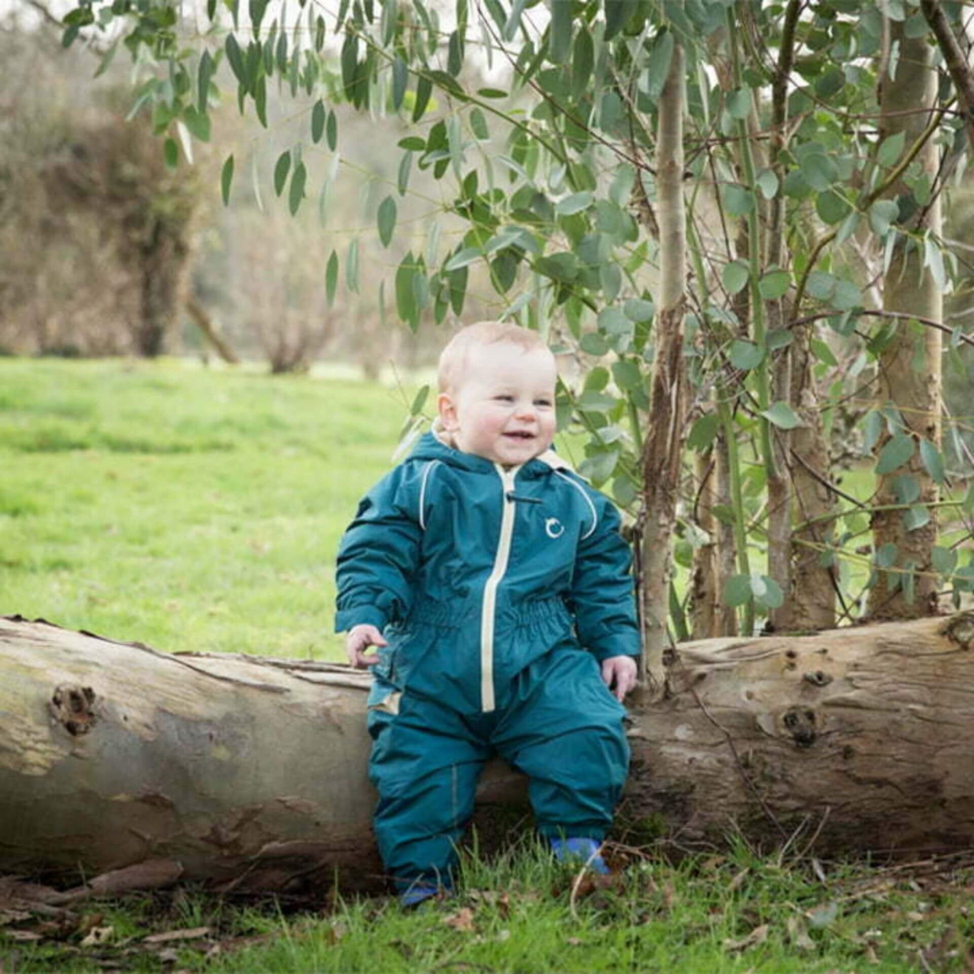 Happy baby wearing a peacock green waterproof fleece-lined suit, sitting on a fallen tree surrounded by greenery in an outdoor setting.