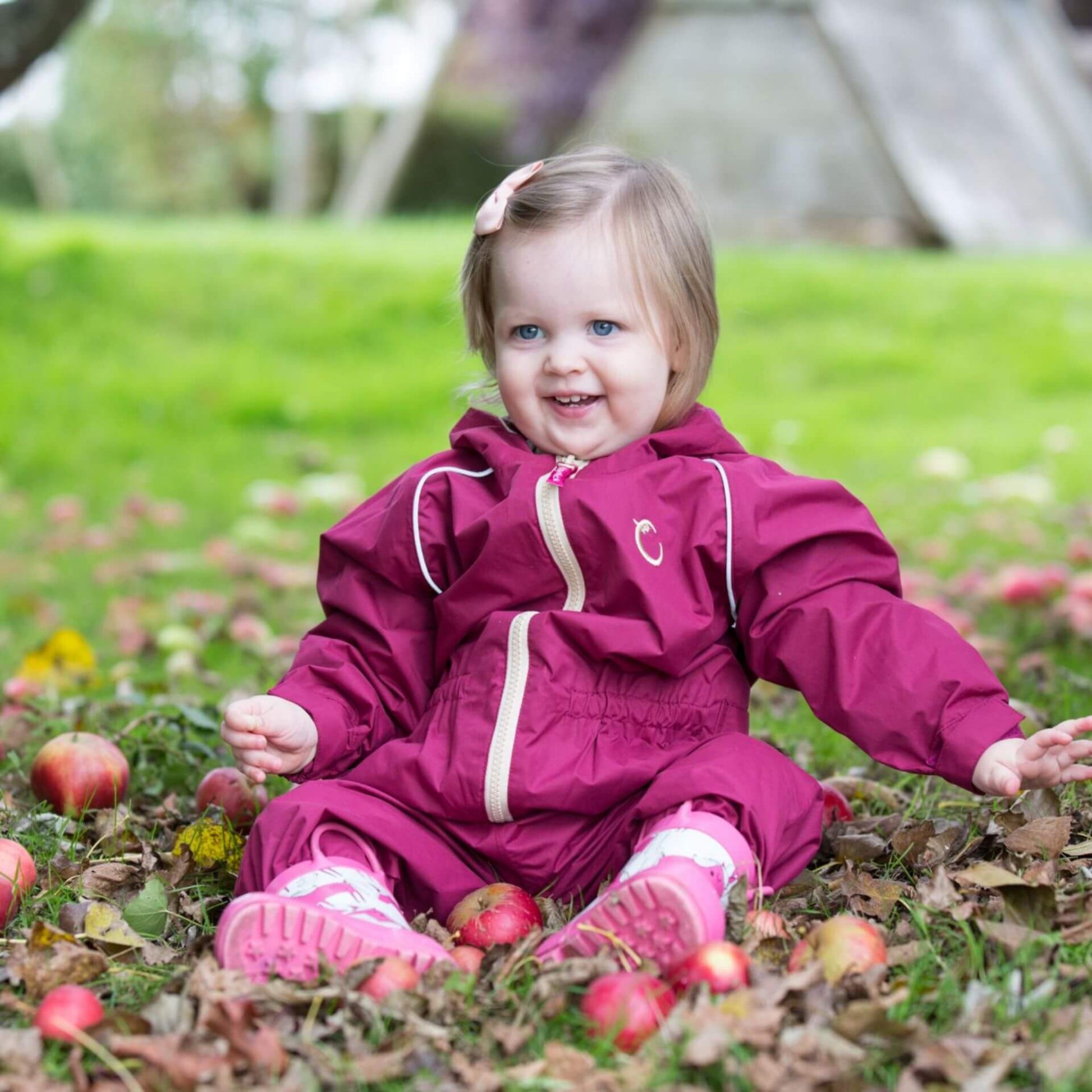 Smiling toddler wearing a raspberry-coloured waterproof fleece-lined suit, sitting on the ground surrounded by fallen apples in an outdoor setting.