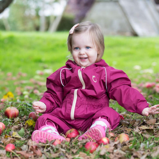 Smiling toddler wearing a raspberry-coloured waterproof fleece-lined suit, sitting on the ground surrounded by fallen apples in an outdoor setting.