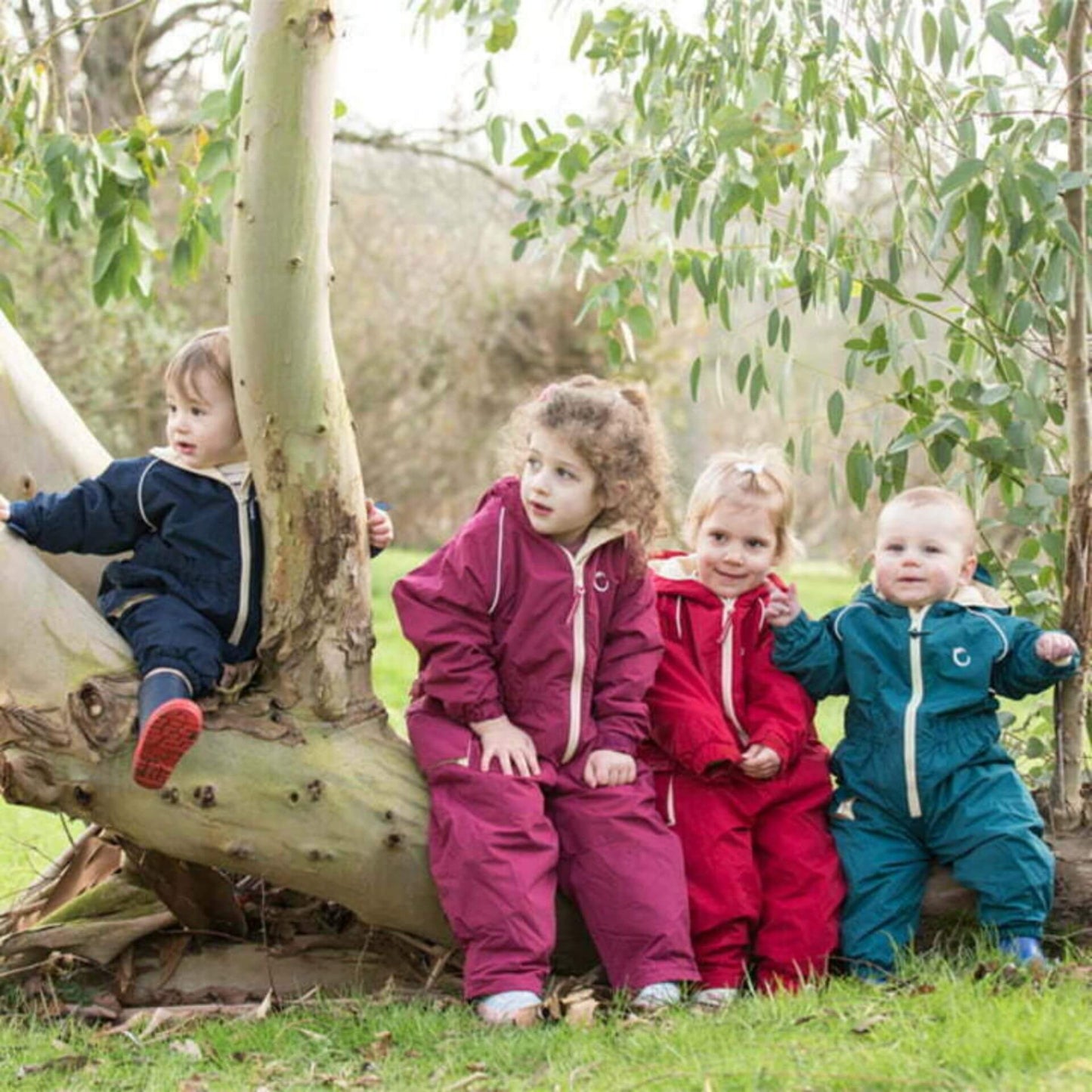 Four children sitting on a fallen tree outdoors, each wearing a different coloured waterproof fleece-lined suit: midnight blue, raspberry, fiesta red, and peacock green.