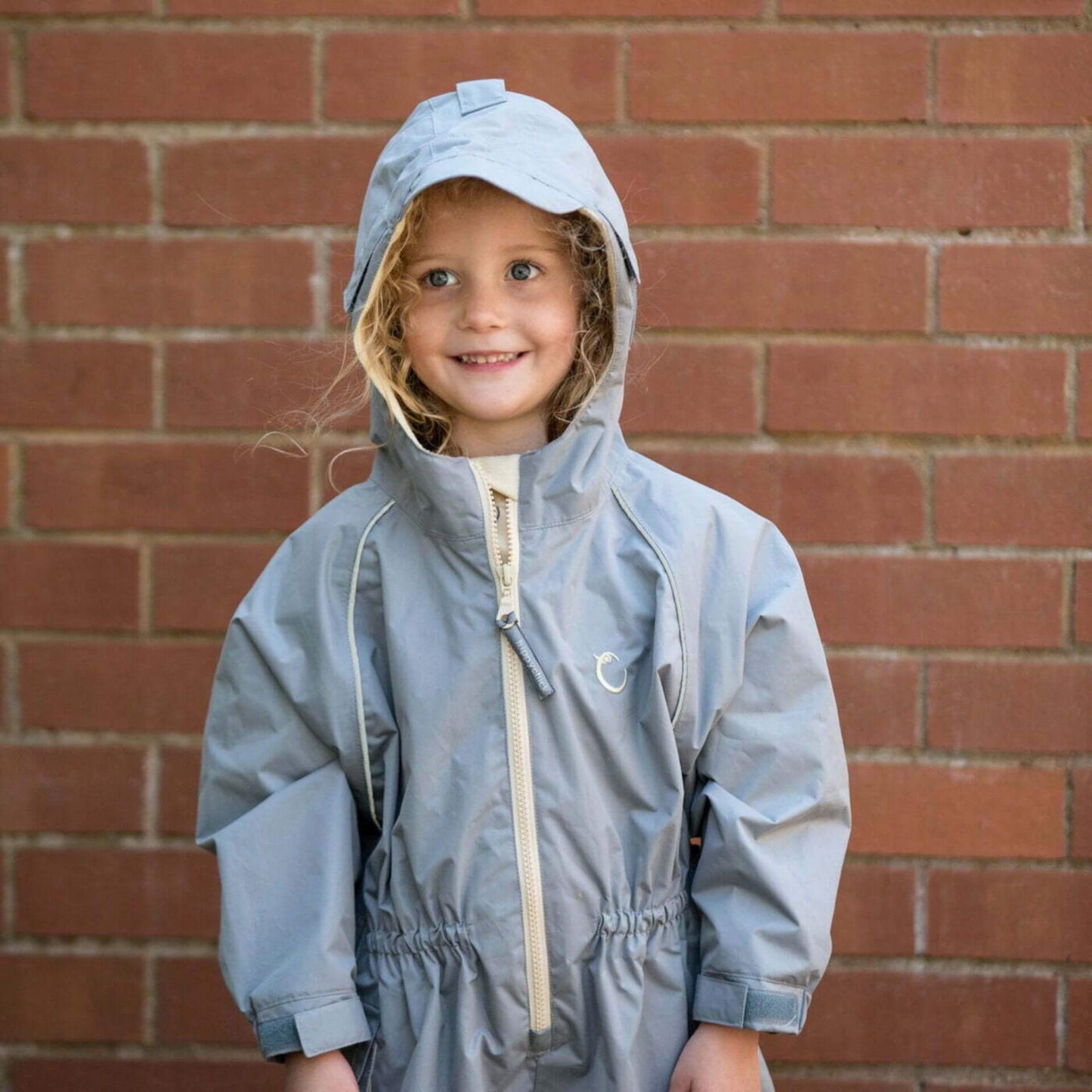 Smiling child wearing a grey waterproof all-in-one suit with hood up, standing against a brick wall background.