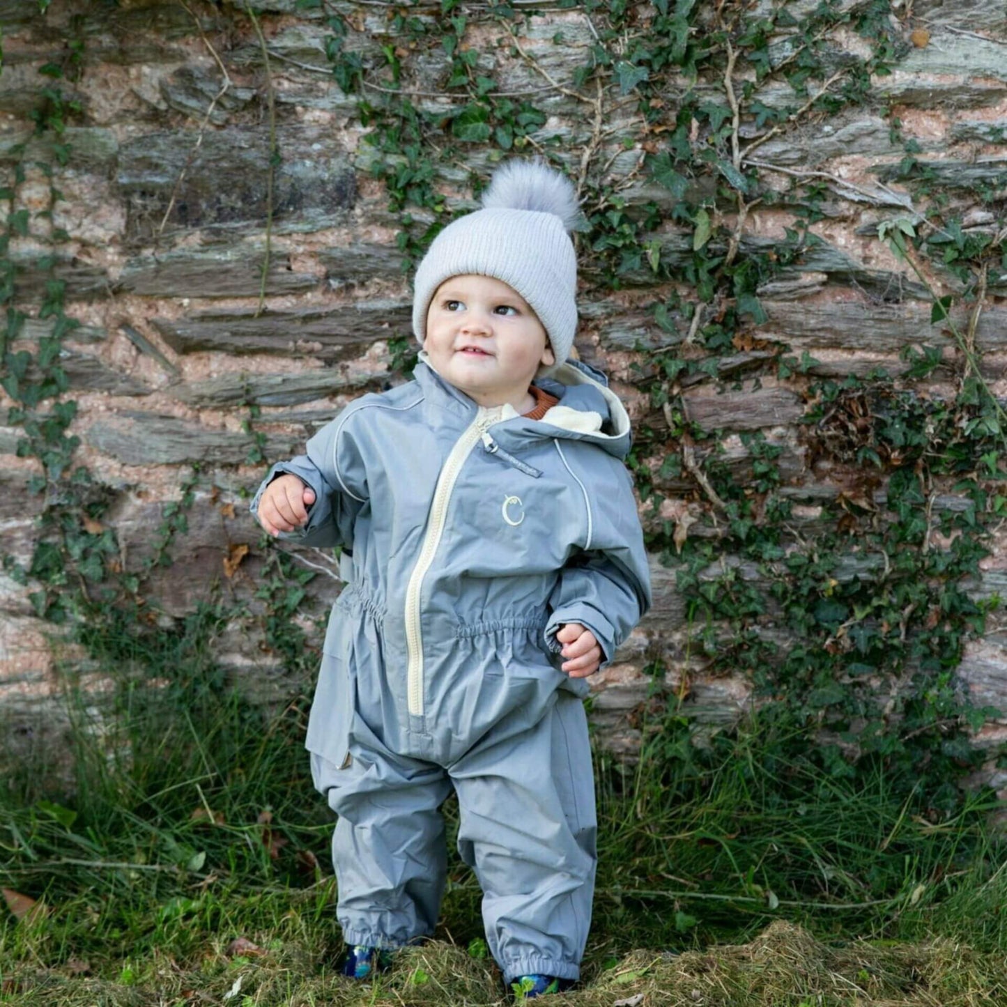 Toddler standing outdoors wearing a grey waterproof all-in-one suit with beige zip and hood, paired with a knitted grey hat with pom-pom.