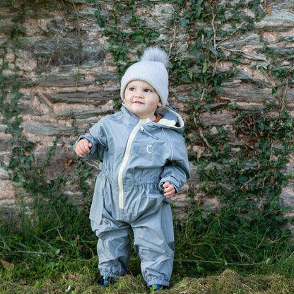 Toddler standing outdoors wearing a grey waterproof all-in-one suit with beige zip and hood, paired with a knitted grey hat with pom-pom.