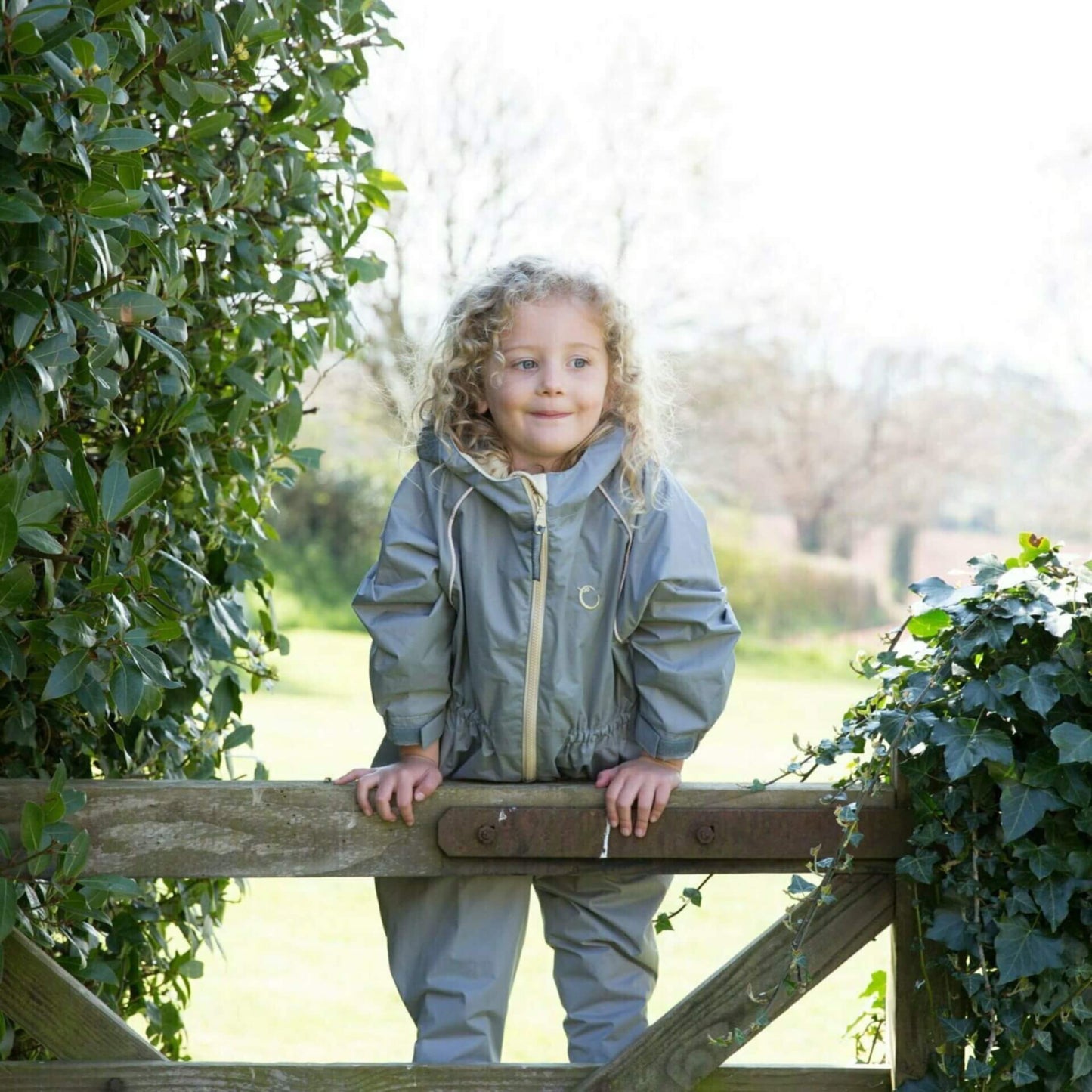 Child leaning on a wooden gate in a grey waterproof all-in-one suit, surrounded by greenery in soft daylight.