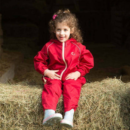 Young child sitting on hay wearing a red waterproof all-in-one suit with hood and beige zip, smiling and dressed for outdoor play.