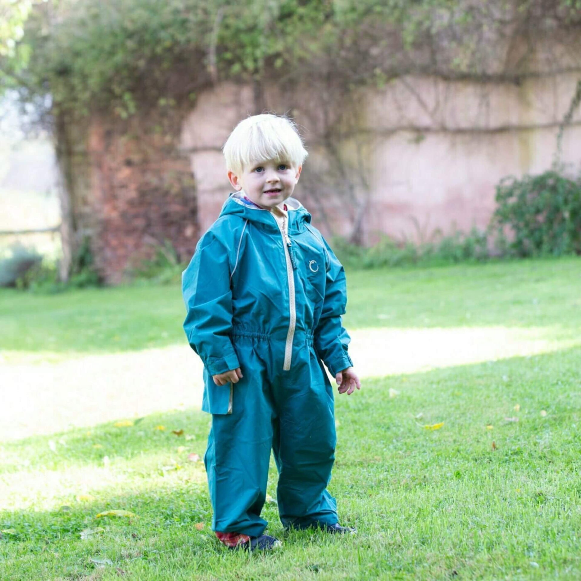Toddler standing on grass wearing a teal-green waterproof all-in-one suit with hood and beige zip, smiling and ready to play outside.