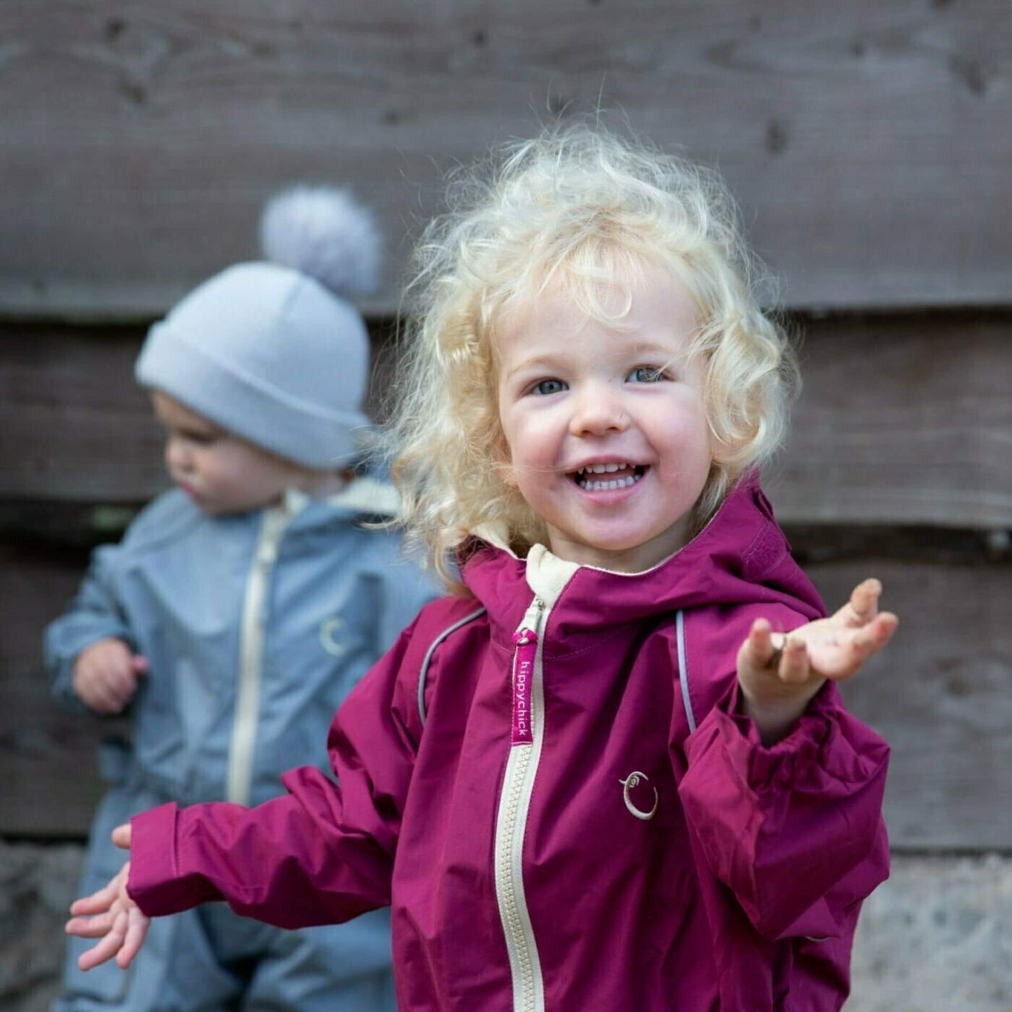 Smiling child wearing a raspberry waterproof all-in-one suit with hood and beige zip, standing outside beside another child in a grey suit.