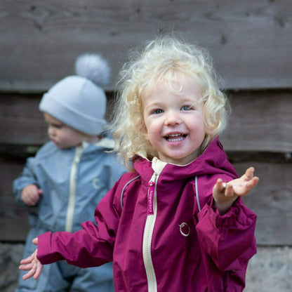 Smiling child wearing a raspberry waterproof all-in-one suit with hood and beige zip, standing outside beside another child in a grey suit.