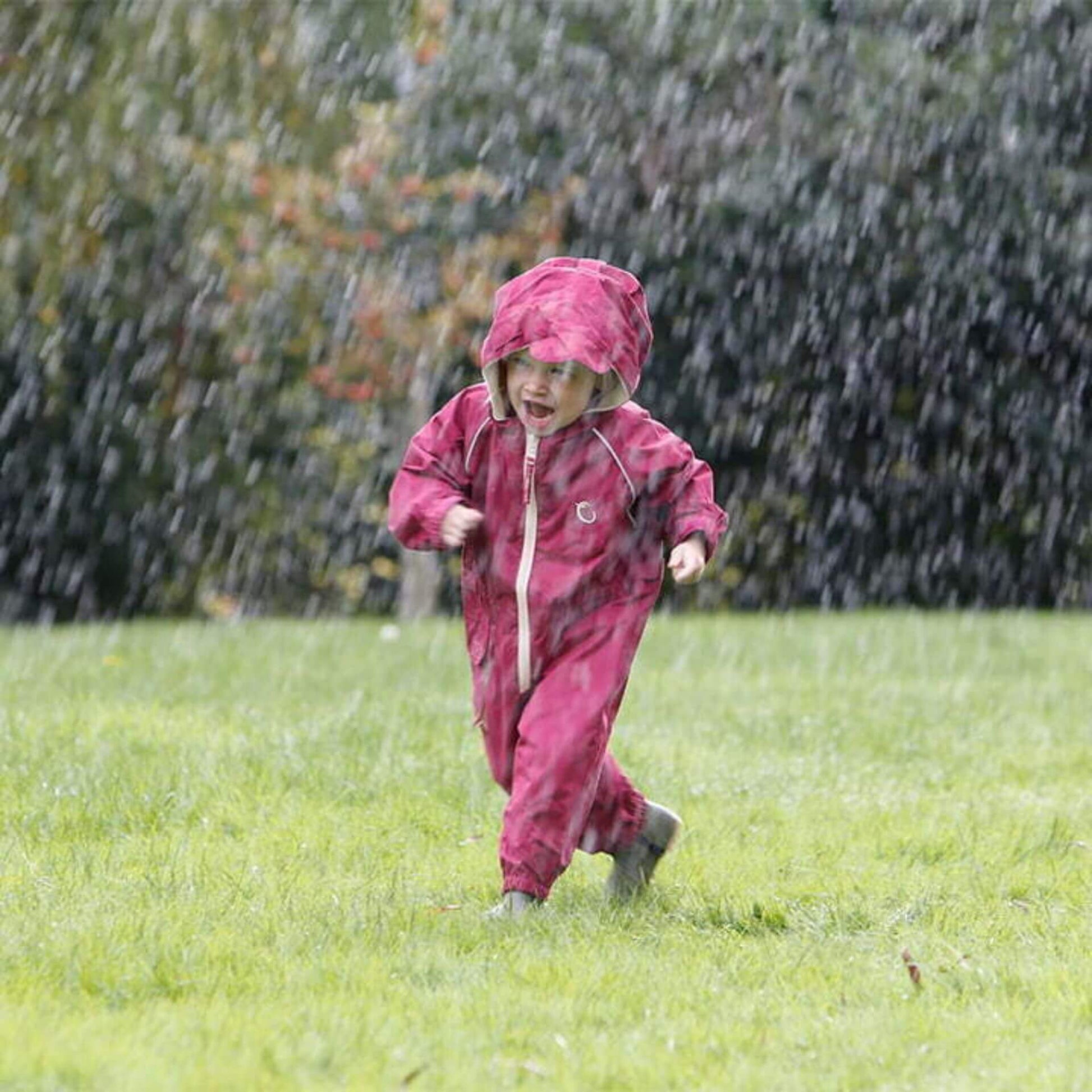 Toddler running through rain wearing a raspberry waterproof all-in-one suit with hood and beige zip, laughing and playing outdoors.