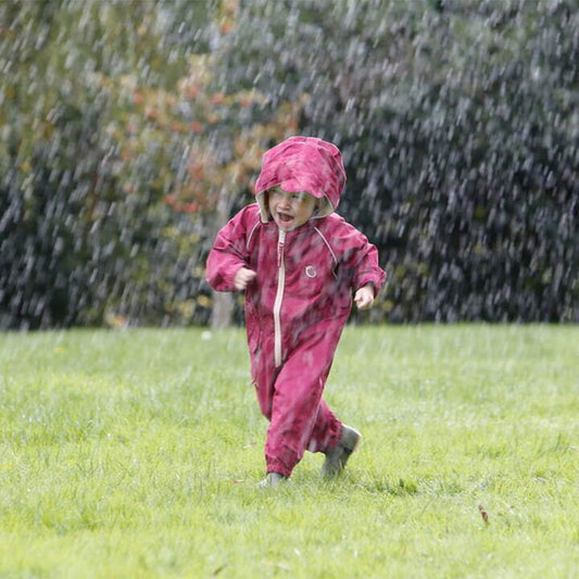 Toddler running through rain wearing a raspberry waterproof all-in-one suit with hood and beige zip, laughing and playing outdoors.