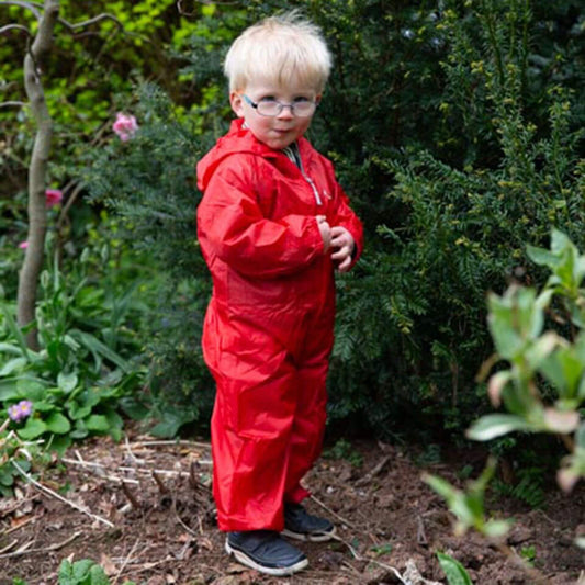 Young child wearing a red waterproof all-in-one suit while standing on soil near bushes outdoors.