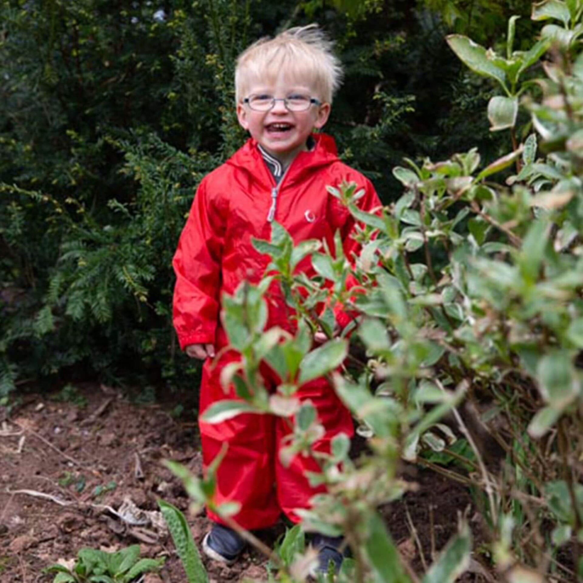 Smiling child in a red waterproof suit with glasses, standing among garden plants and greenery.