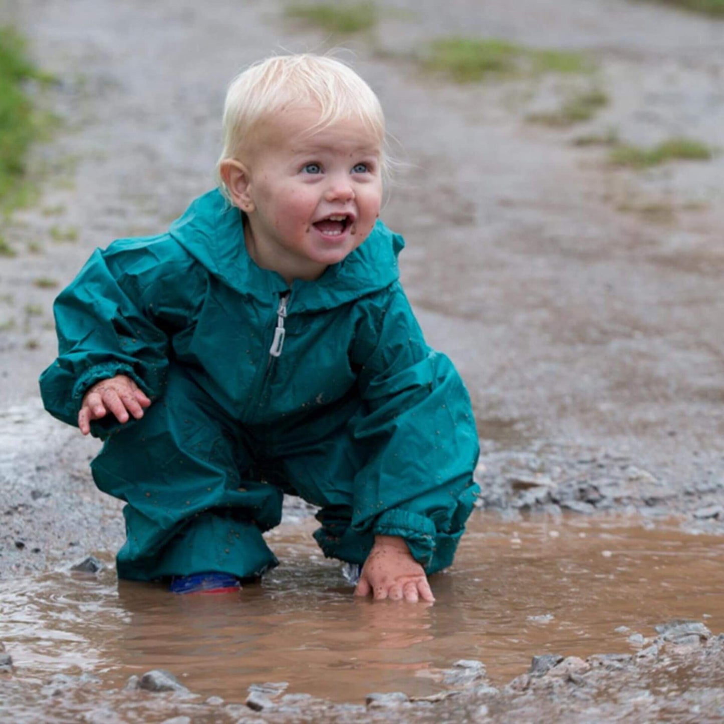 Toddler crouching in a muddy puddle wearing a green waterproof packasuit with sleeves and legs gathered at the cuffs.