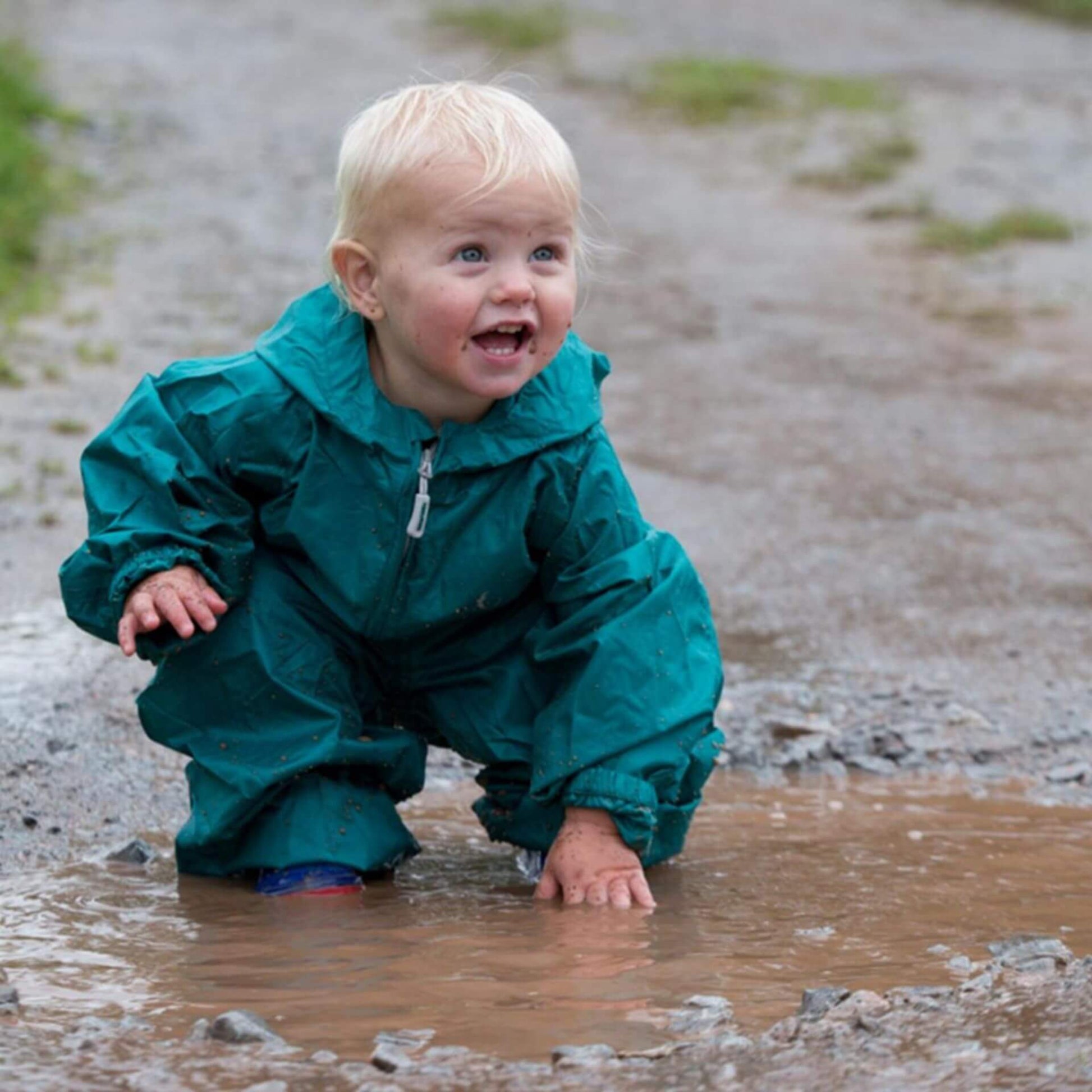 Toddler crouching in a muddy puddle wearing a green waterproof packasuit with sleeves and legs gathered at the cuffs.