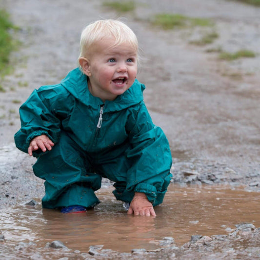 Toddler crouching in a muddy puddle wearing a green waterproof packasuit with sleeves and legs gathered at the cuffs.