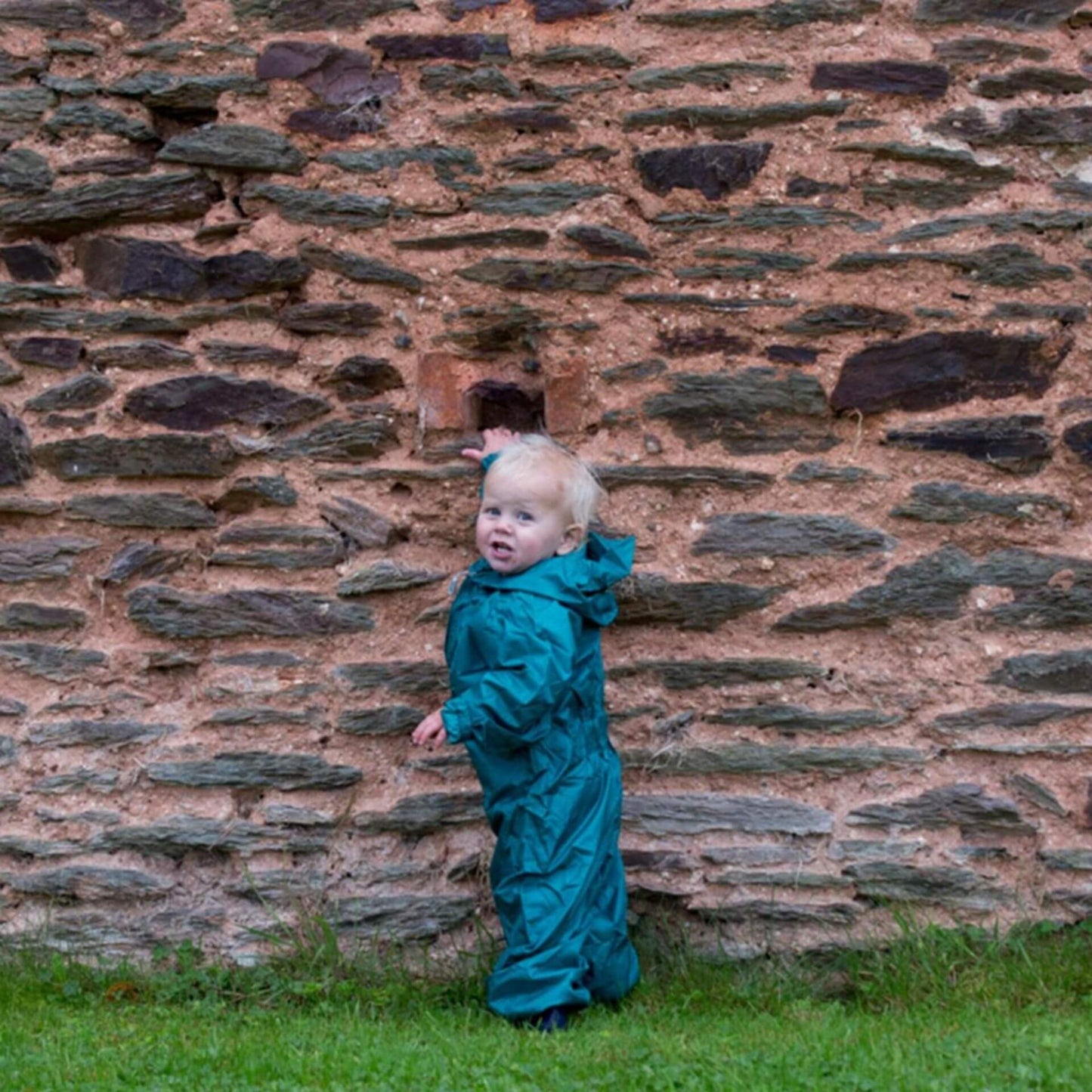 Child standing outdoors against a stone wall dressed in a bottle green waterproof suit with hood up.