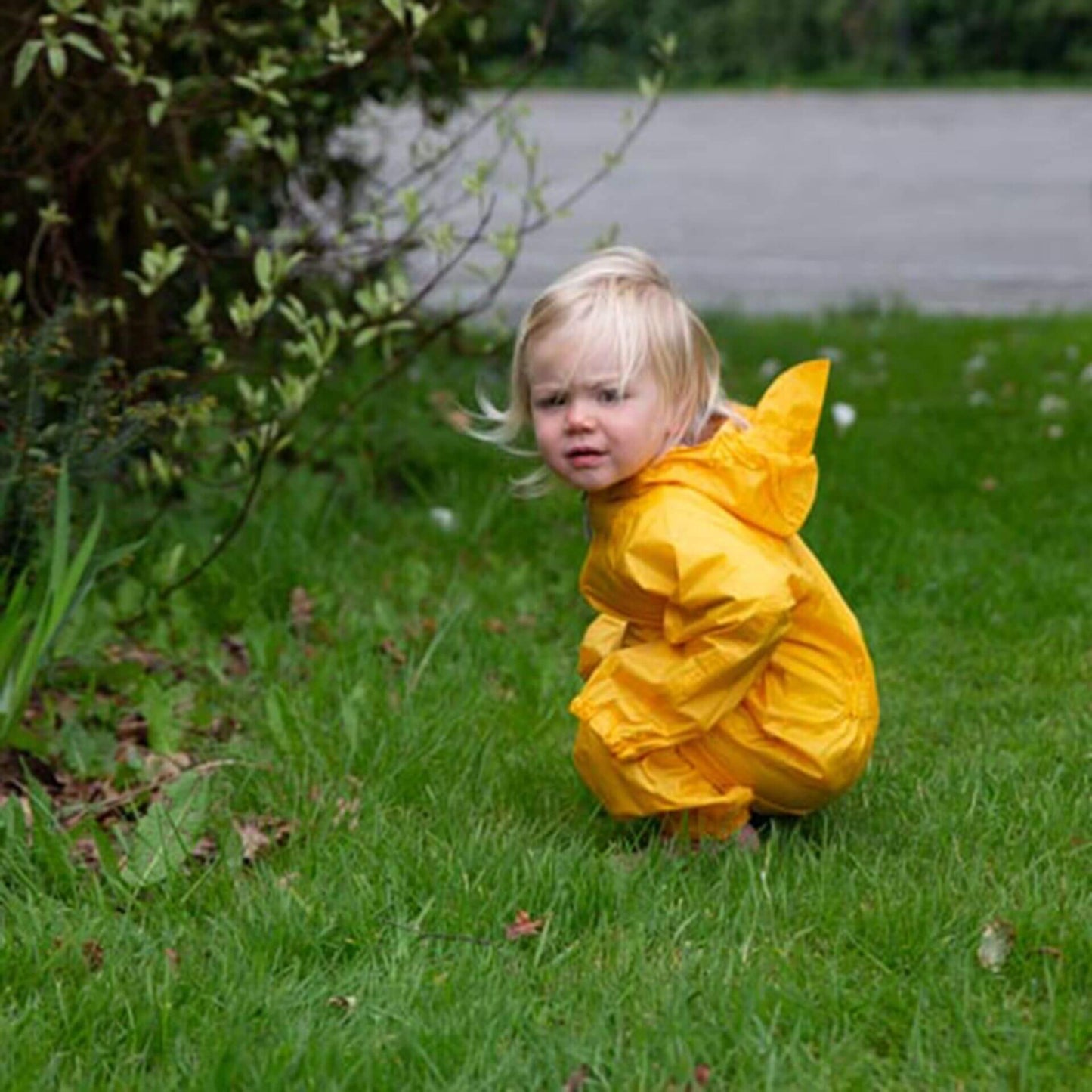 Child crouching on grass in a yellow waterproof packasuit with hood up, surrounded by garden plants.