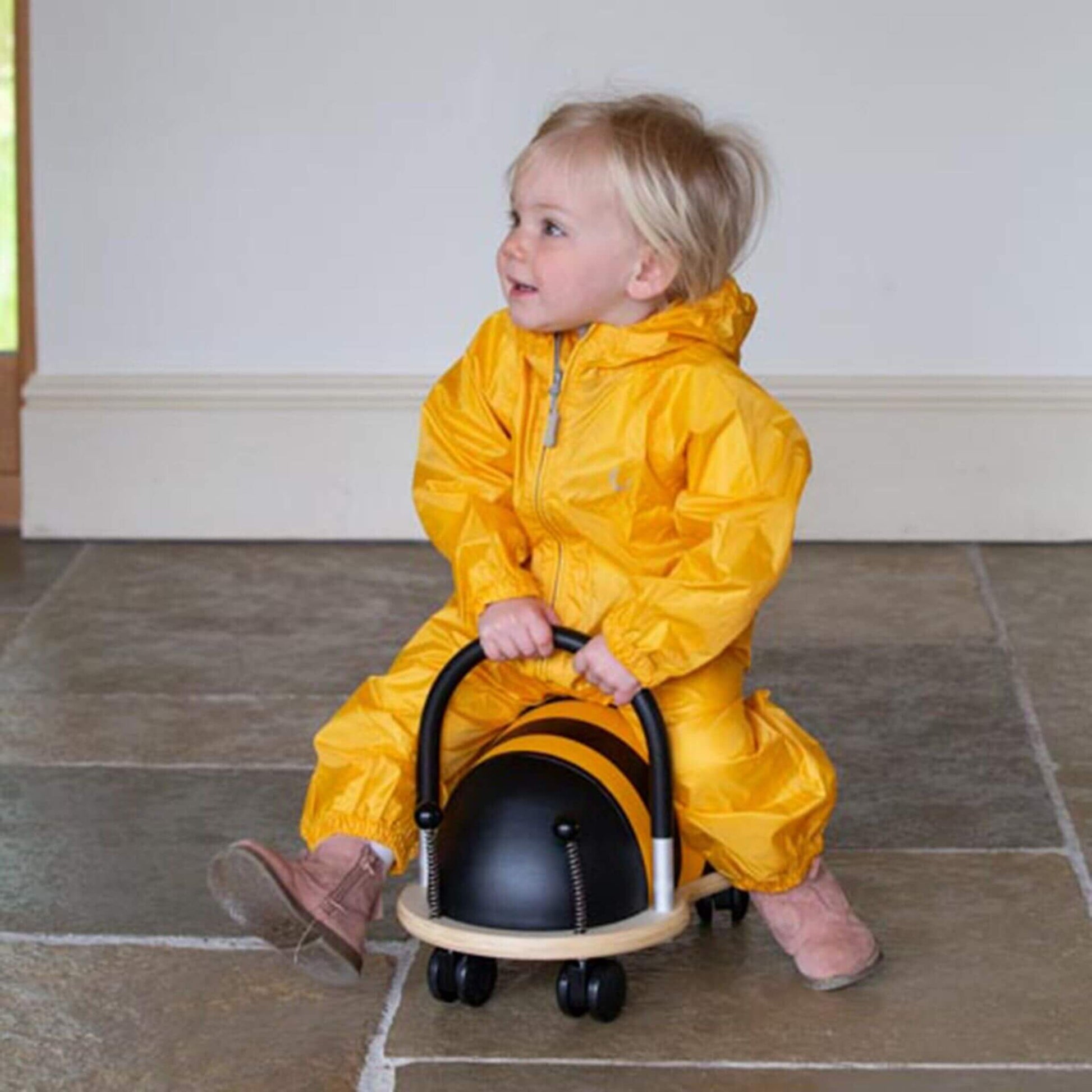 Toddler sitting indoors on a ride-on toy bee while wearing a yellow waterproof packasuit, looking to the side with a smile.