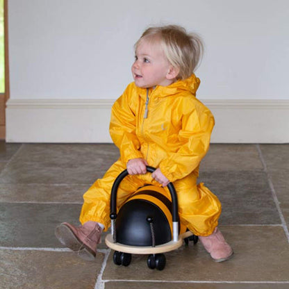 Toddler sitting indoors on a ride-on toy bee while wearing a yellow waterproof packasuit, looking to the side with a smile.
