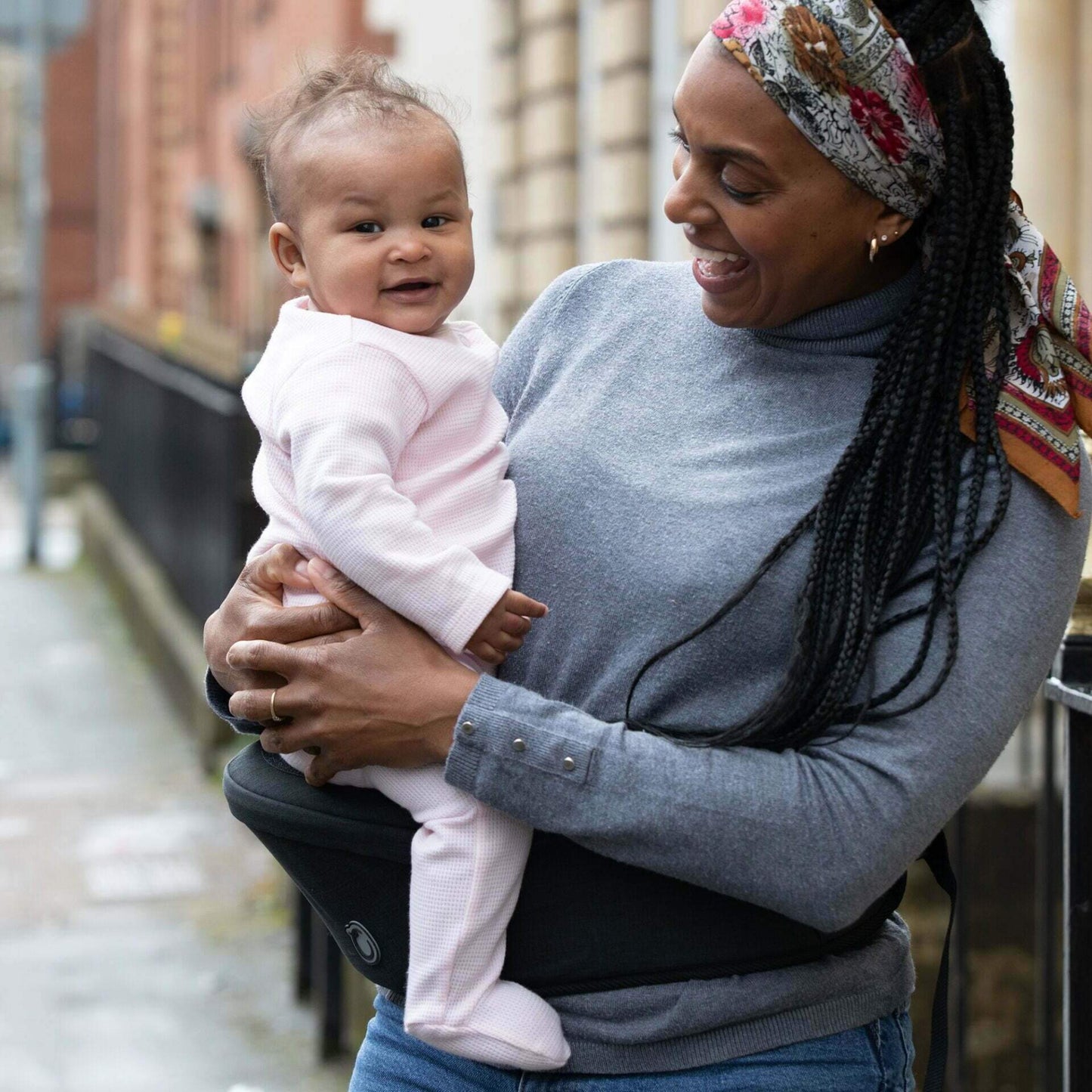 Smiling mother wearing the black Hipseat carrier around her waist, comfortably holding her baby who is dressed in a pale pink sleepsuit.