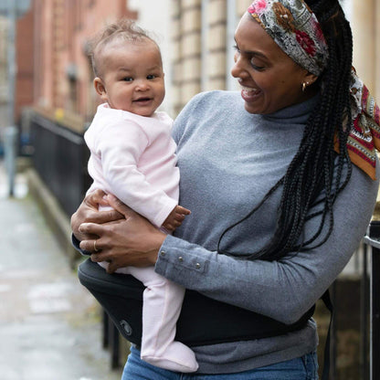 Smiling mother wearing the black Hipseat carrier around her waist, comfortably holding her baby who is dressed in a pale pink sleepsuit.
