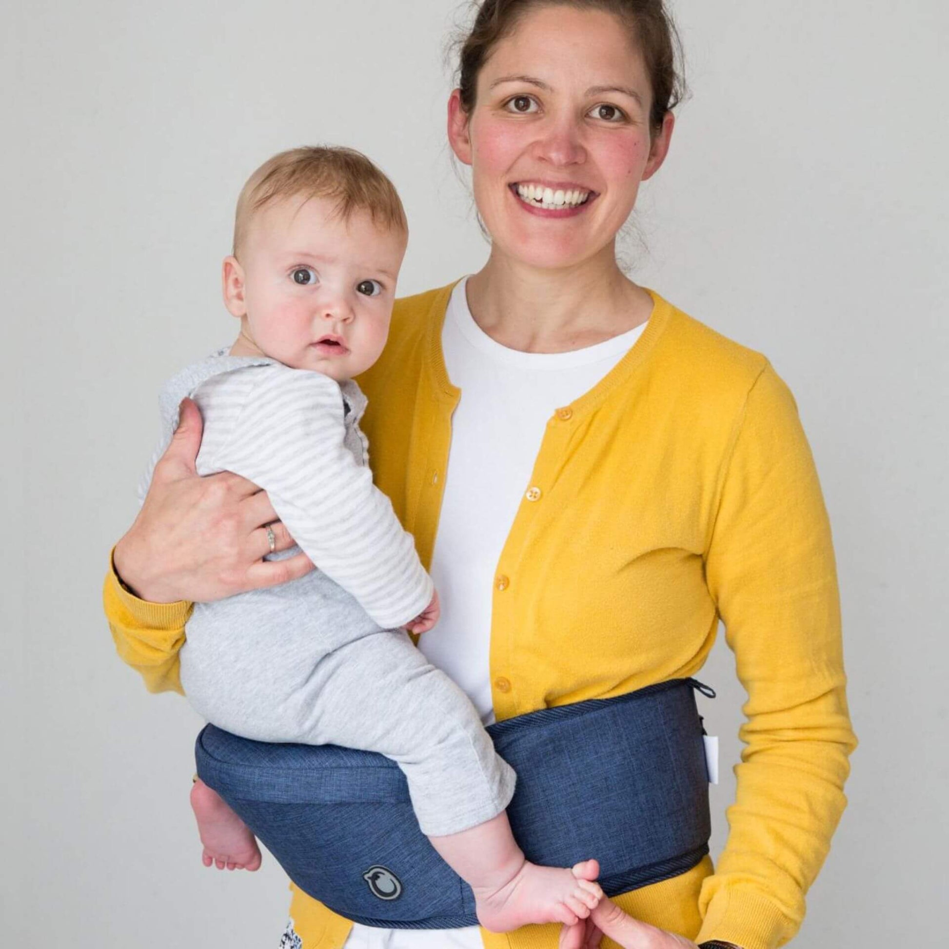 A smiling mother wearing a yellow cardigan holds her baby securely while using a blue Hippychick Hipseat carrier around her waist.