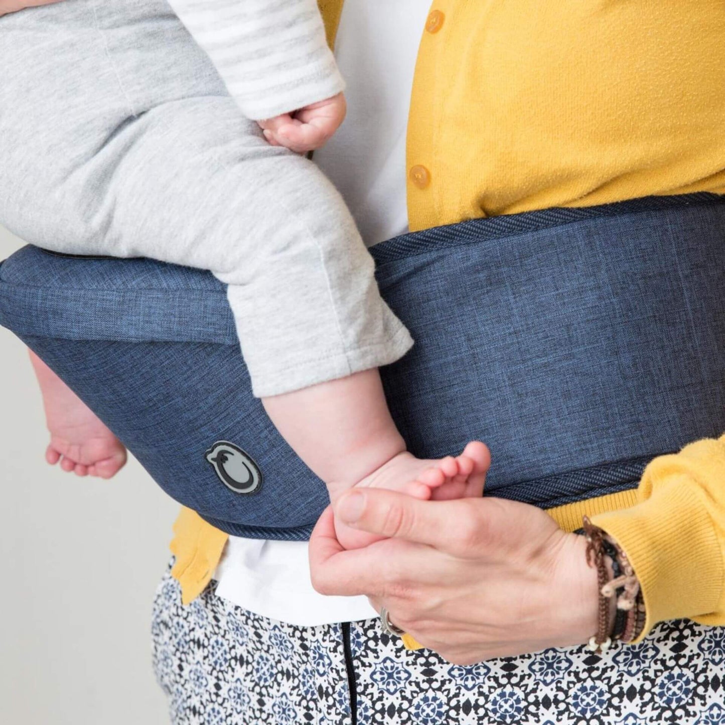 Close-up view of a baby’s legs resting on a blue denim baby Hipseat carrier, showing the padded seat and waistband detail in use.
