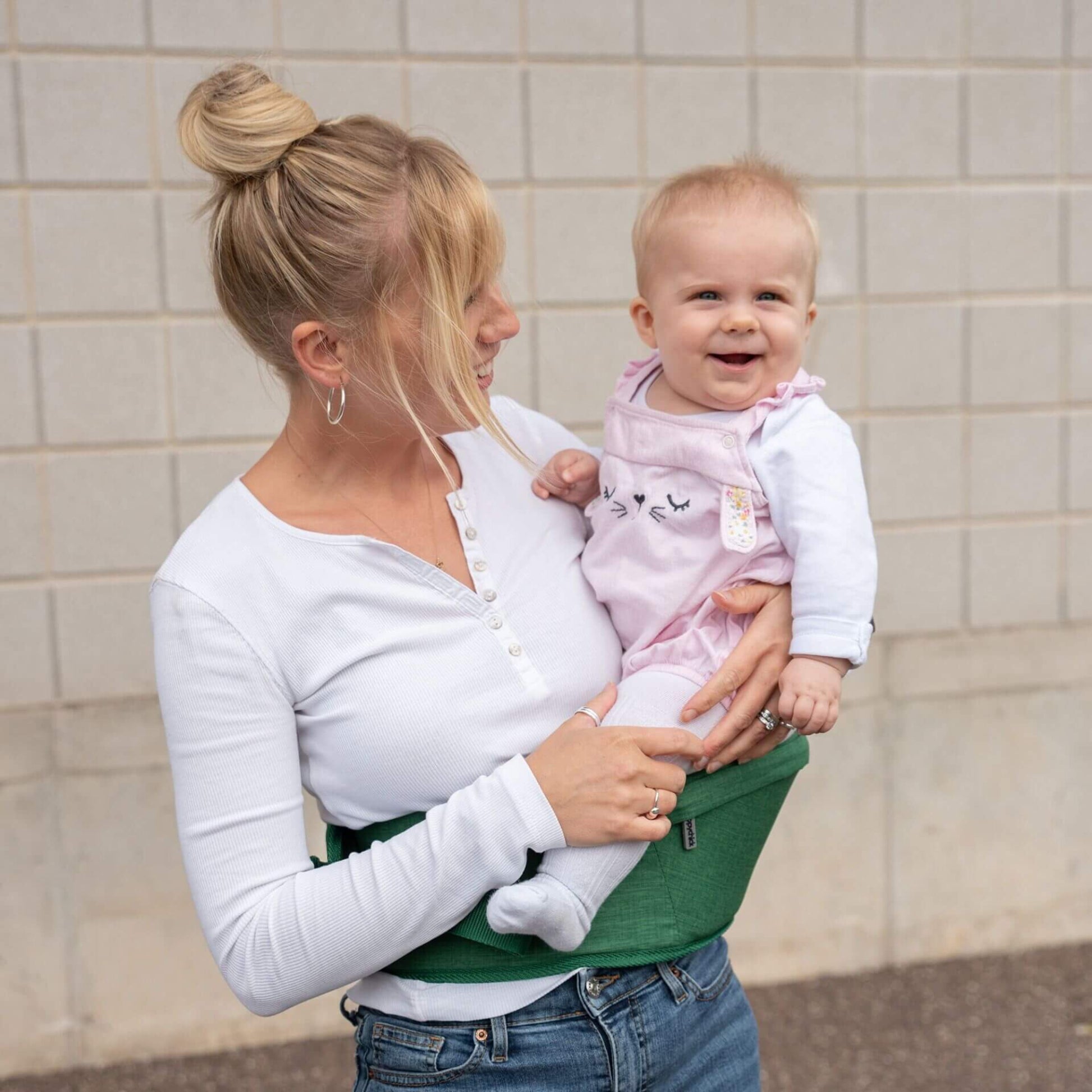 Mother carrying smiling baby in a white top and pink dungarees using a green hipseat outdoors.