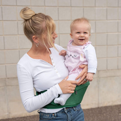 Mother carrying smiling baby in a white top and pink dungarees using a green hipseat outdoors.