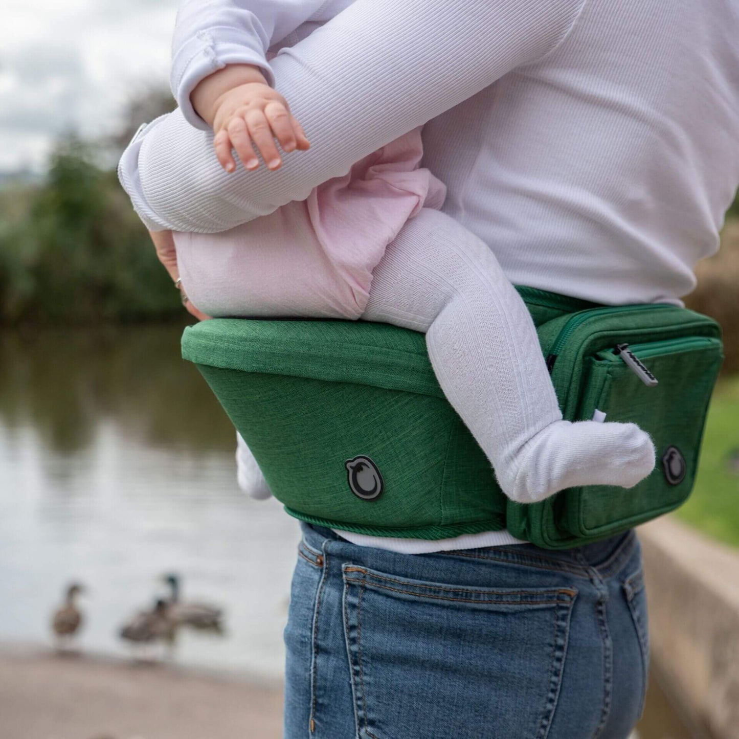 Close-up side view of a baby sitting securely on padded green hipseat with zipped storage pouch.