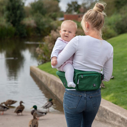 Rear view of mother standing by a pond with a baby sitting on a green hipseat, ducks in the background.