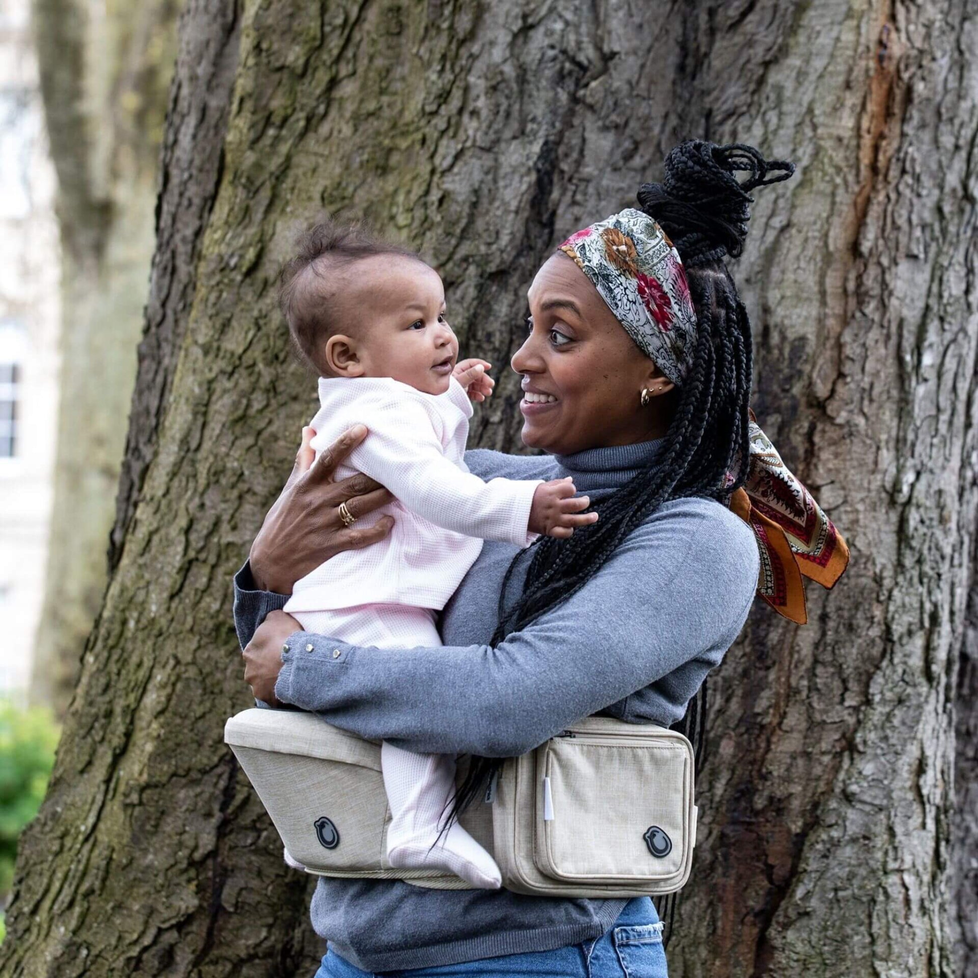 Smiling mother outdoors using the Hipseat Denim Oatmeal to comfortably carry her baby while standing by a large tree.