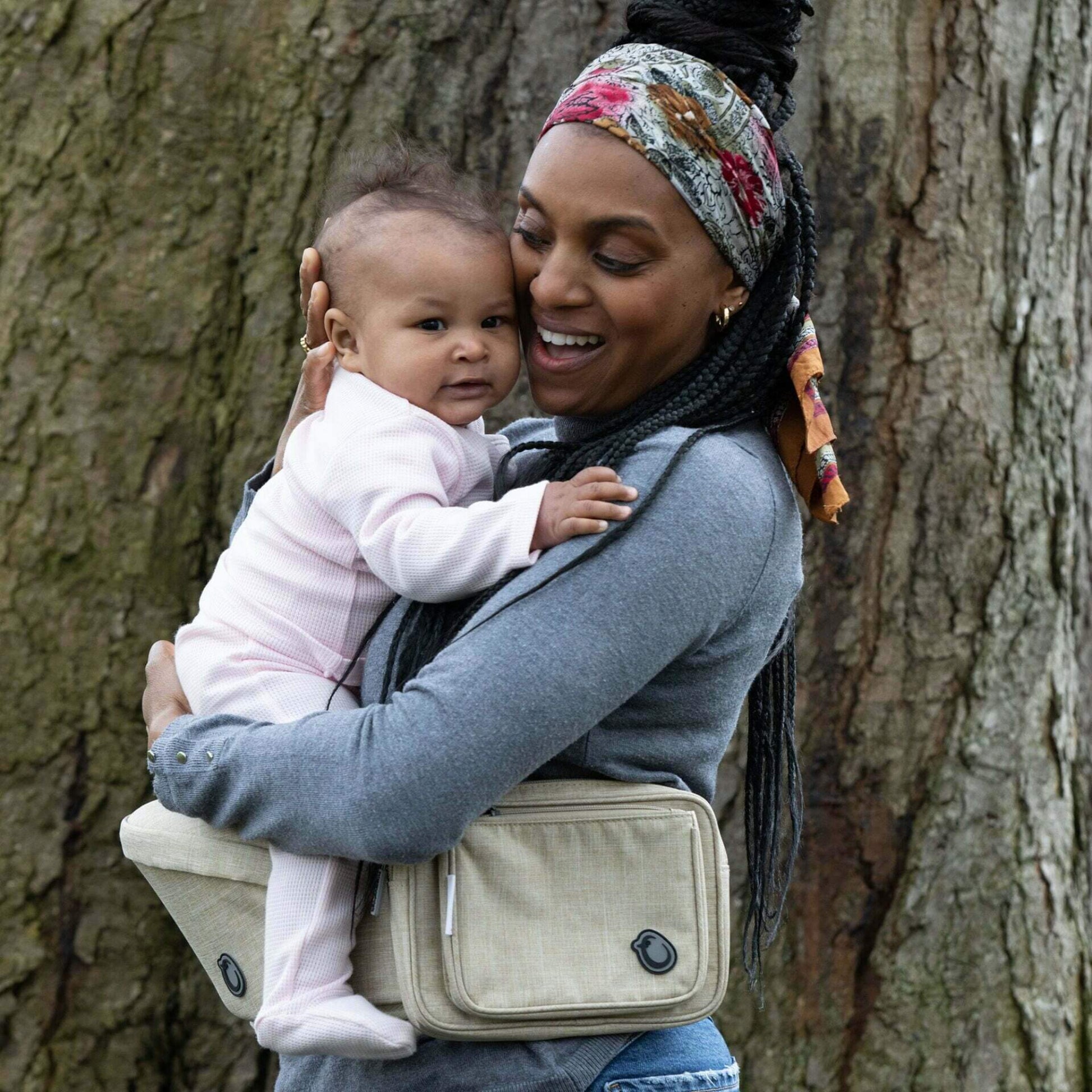 Mother holding her baby close while using the Hipseat Denim Oatmeal, both looking content in an outdoor woodland setting.