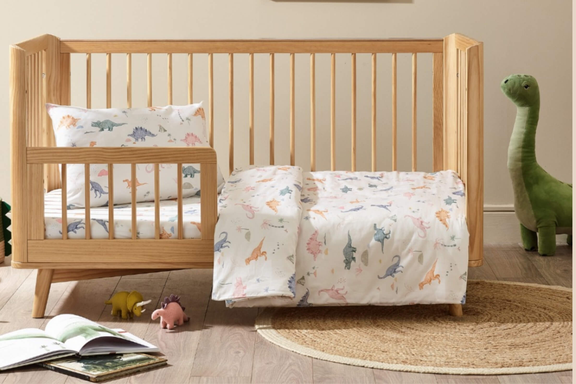 A wooden toddler bed with vertical slats, dressed in dinosaur-print bedding, set on a wooden floor with small toy dinosaurs, an open book, a woven rug, and a soft dinosaur toy beside the bed.