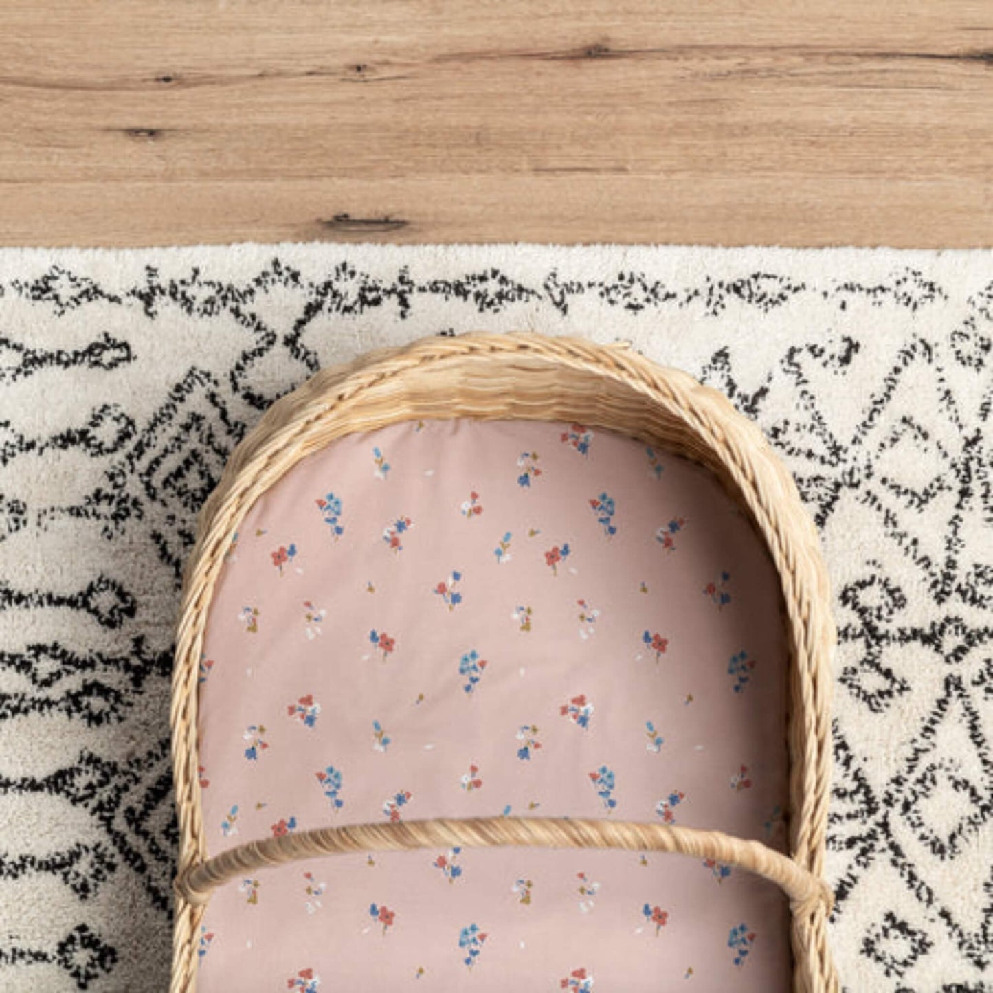A woven Moses basket mattress covered with a soft pink fitted sheet, featuring a delicate floral print, photographed from above on a patterned rug.