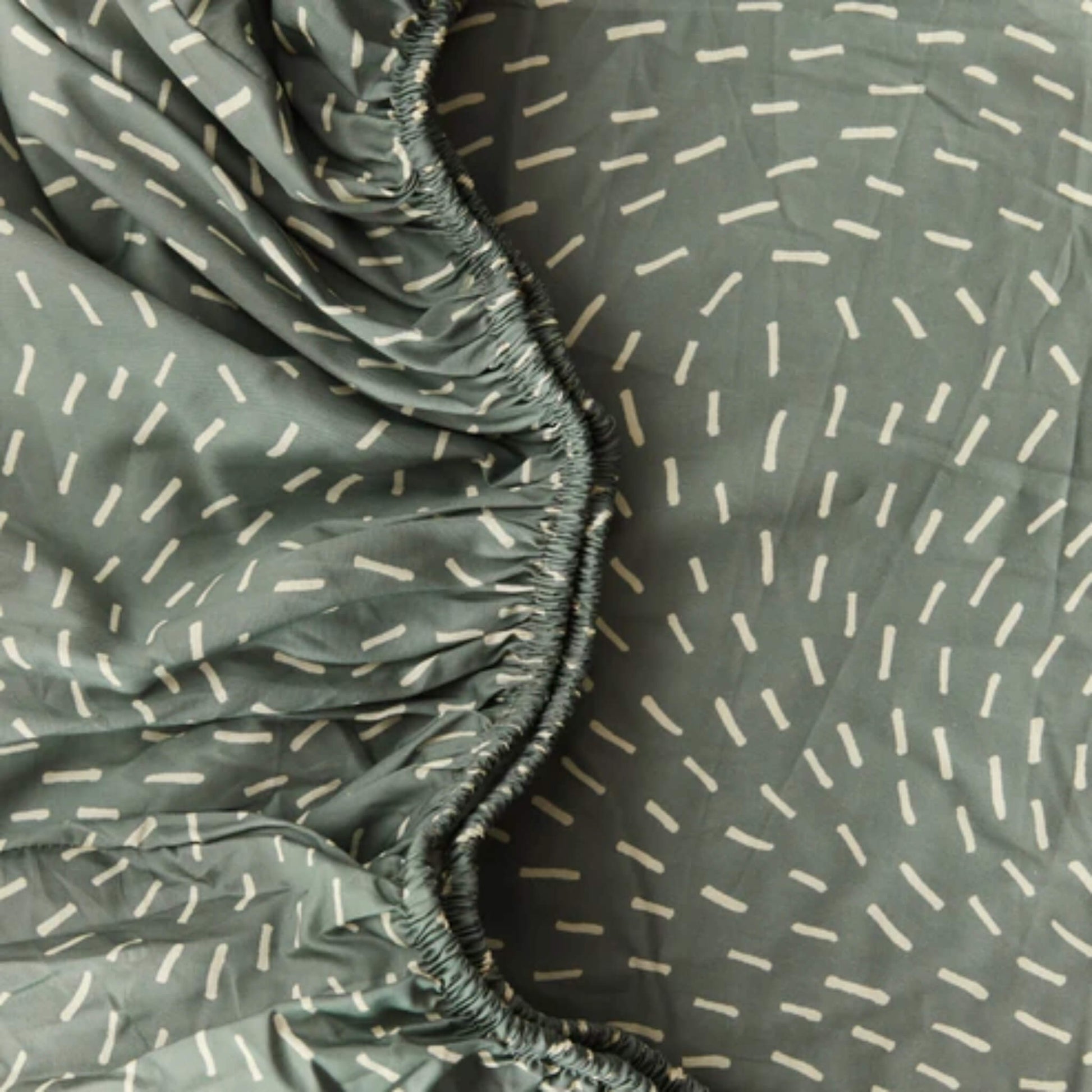 Close-up of organic cotton fitted sheet fabric in sage green with white dash pattern, showing smooth texture.