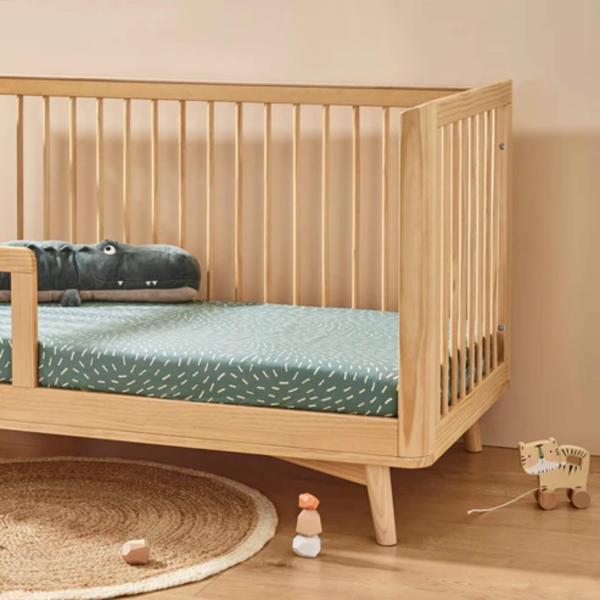 Organic cotton fitted sheet in Dashes print shown on a wooden cot bed with sage green mattress cover and neutral nursery setting.