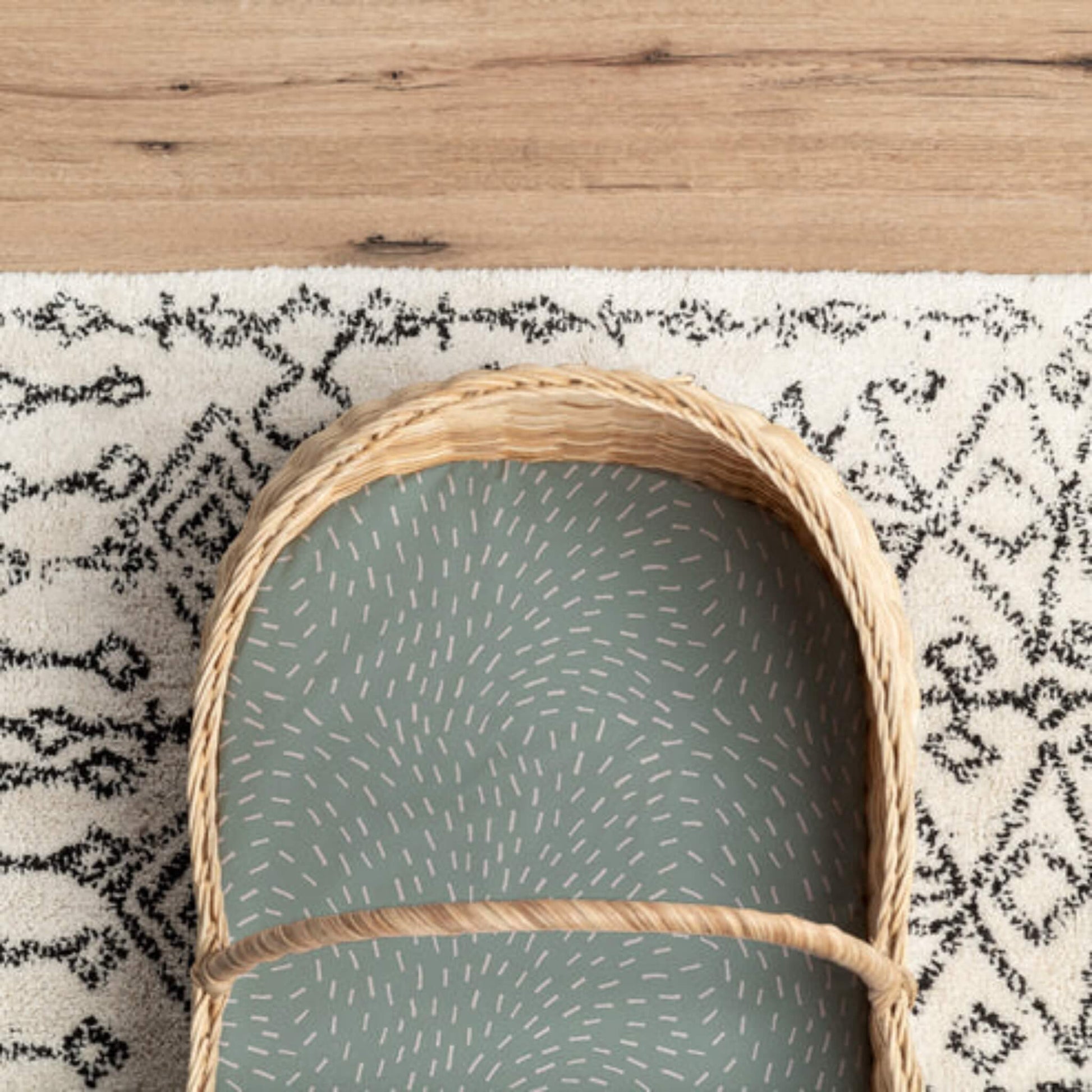 Fitted sheet in sage green Dashes print shown inside a Moses basket with woven frame and patterned rug beneath.