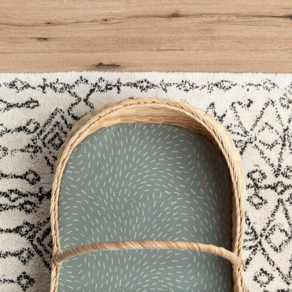 Fitted sheet in sage green Dashes print shown inside a Moses basket with woven frame and patterned rug beneath.