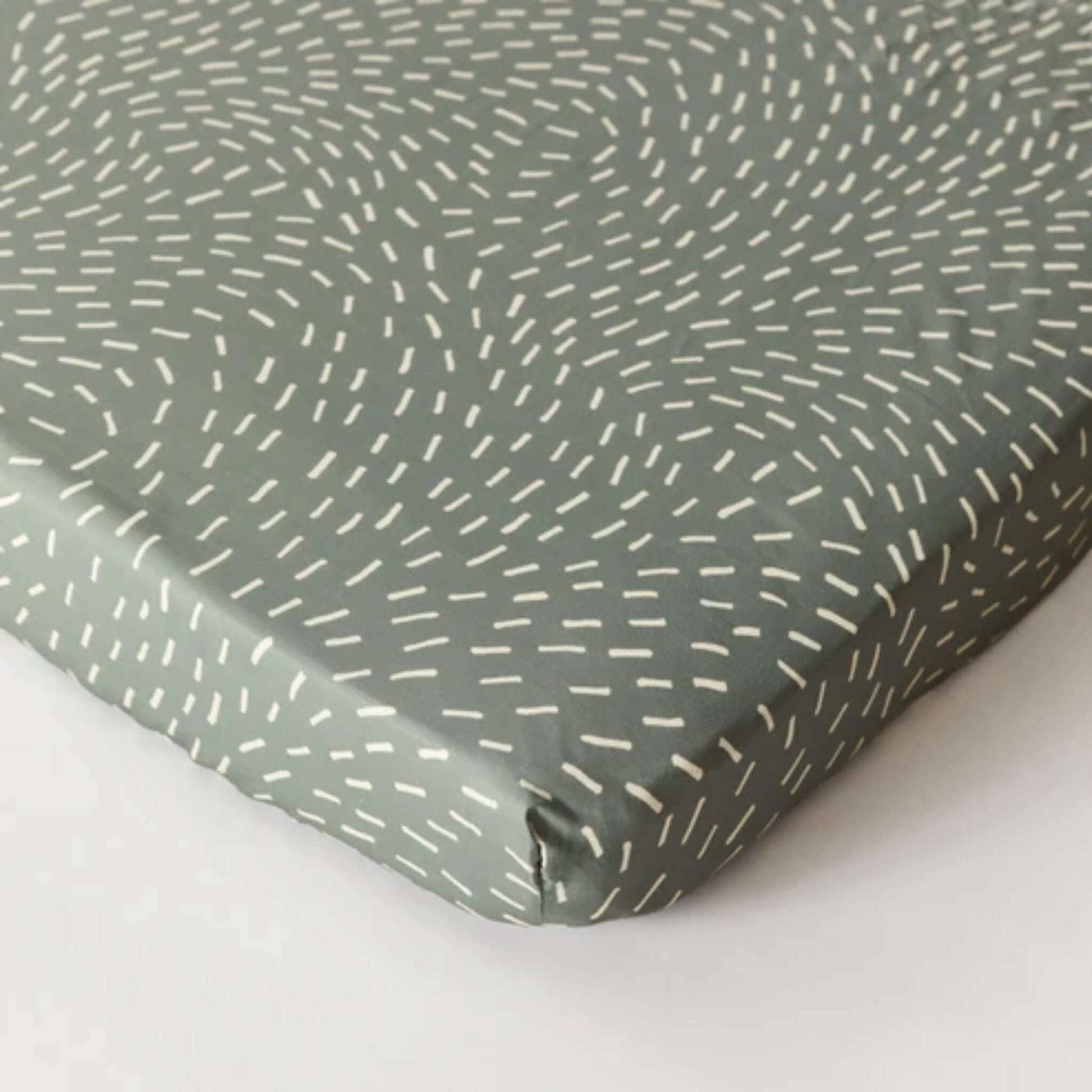 Kabode organic cotton fitted sheet in Dashes print, shown fitted over a mattress corner in soft sage green with white dash pattern.
