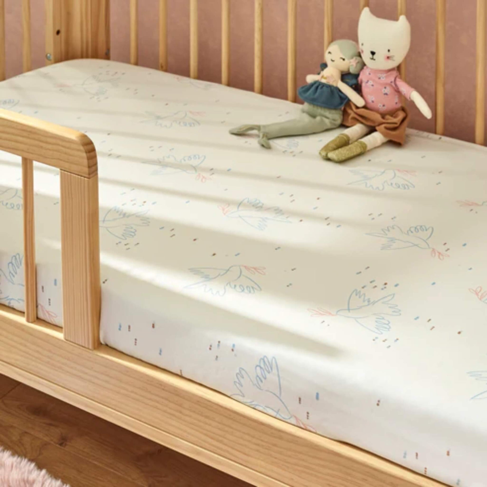 A cot bed dressed with a cream fitted sheet featuring delicate bird illustrations, styled with soft toys on a wooden frame.