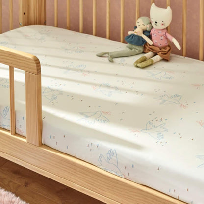 A cot bed dressed with a cream fitted sheet featuring delicate bird illustrations, styled with soft toys on a wooden frame.